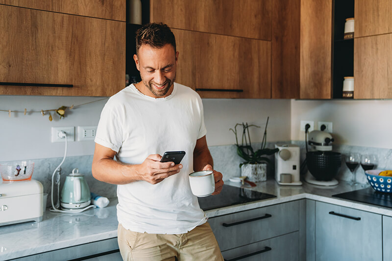 A man in a white shirt holds a mug and smartphone, smiling in a modern kitchen with wooden cabinets and appliances on the counter.