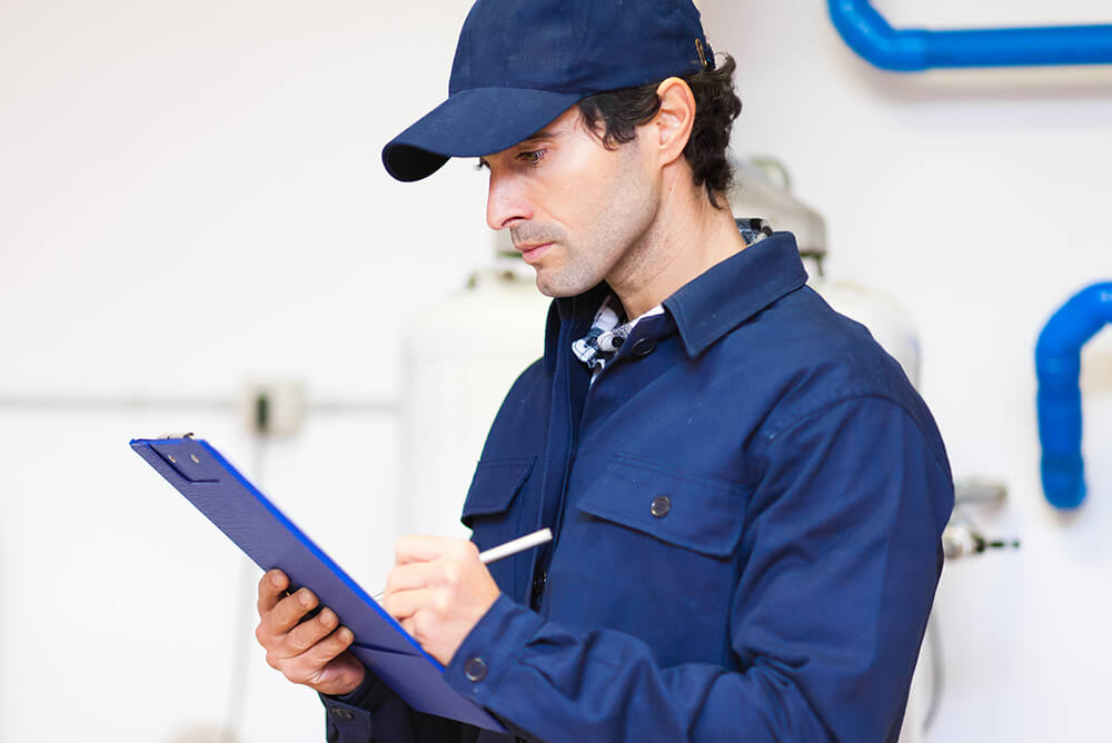 A man in a blue uniform writes on a clipboard. He stands in a room with visible pipes and industrial equipment in the background.