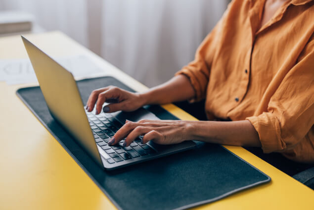 A person types on a laptop placed on a yellow desk with a black desk mat, near a window with white curtains.