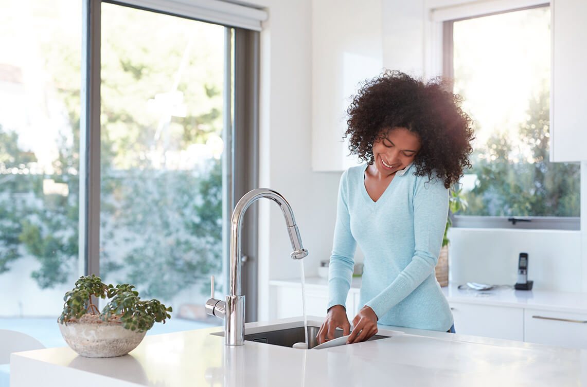 A person washes dishes at a kitchen sink, smiling, with natural light entering through large windows, and a plant on the counter.