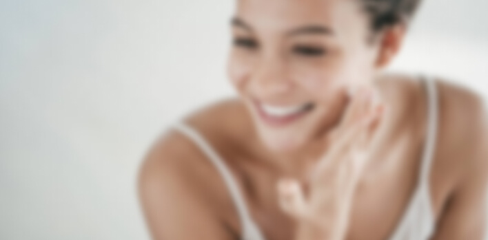 A smiling person touches their cheek, wearing a light camisole, against a soft, blurred background, suggesting a relaxed and gentle atmosphere.